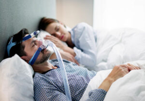 man laying in bed with cpap machine and wife sleeping next to him.